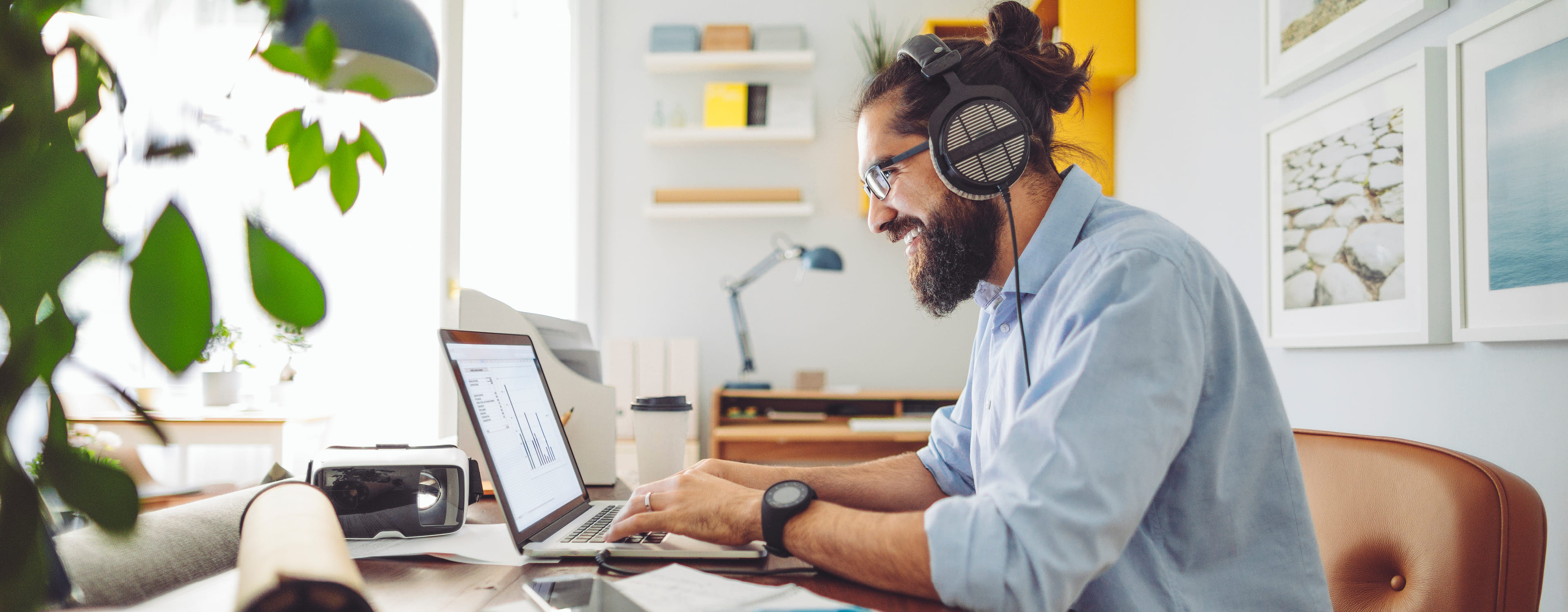 Person working on a laptop at a desk with papers, a coffee cup, and electronic devices. Background includes shelves with books, decorative items, and framed pictures. Face is pixelated for privacy.