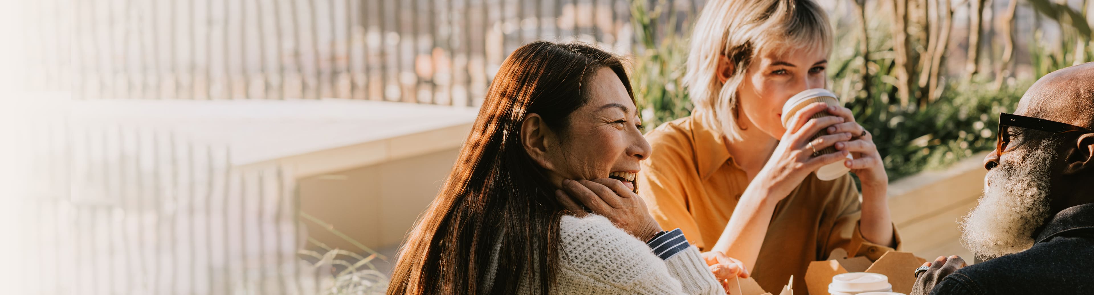 Three people sitting at an outdoor table, enjoying food and drinks while engaged in conversation. Surrounded by greenery with a blurred natural background, suggesting a casual dining or social gathering setting.