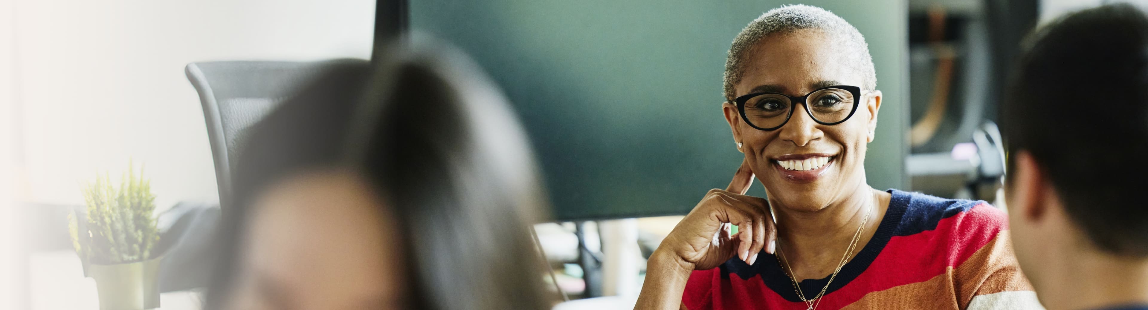 Three people sitting and conversing indoors. The central person wears a colorful striped shirt in red, orange, and blue, along with a necklace. Faces are blurred for privacy.
