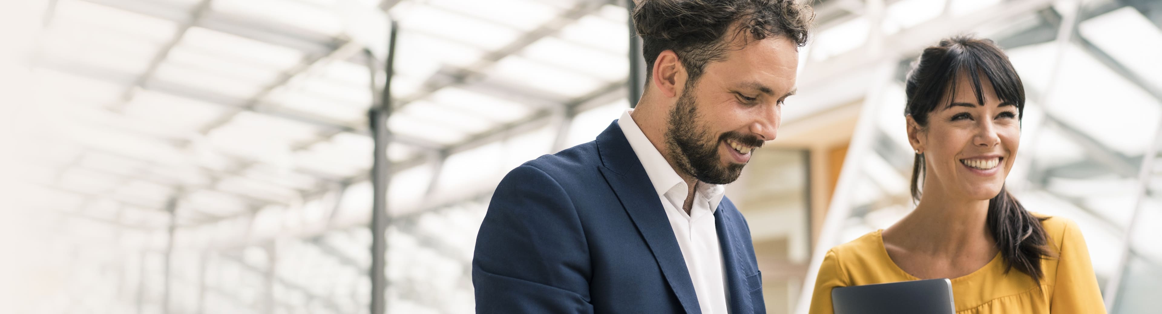 Two people standing close together in a modern indoor space with a glass ceiling and metal framework. One wears a dark blue suit with a white shirt, and the other wears a yellow top. Faces are blurred for privacy.