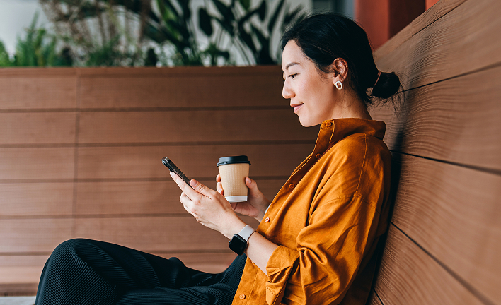 Person in an orange shirt sitting on a wooden bench, holding a smartphone and a takeaway coffee cup. Wearing a smartwatch, with plants and wooden paneling in the background. Face is blurred for privacy.
